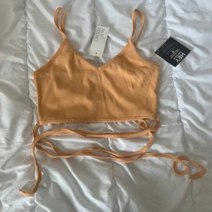 Ribbad croppad linne från NLY Trend - Supersnygg croppad linne i ljus orange från NLY Trend. Toppen har smala axelband, ribbad struktur och långa knytband som kan snurras runt midjan för en trendig look. Perfekt till sommarens outfits och enkel att matcha med jeans eller kjol
