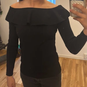Svart offshoulder topp med volang - Säljer en svart offshoulder topp med lång ärm och bred volang runt hela axelpartiet. Toppen har en tight passform och är tillverkad i ett mjukt, stretchigt material som sitter snyggt på kroppen. Perfekt för dig som gillar stilrena och trendiga plagg. Priset kan diskuteras.