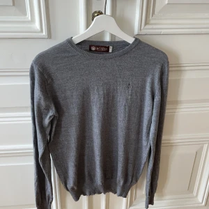 Grå stickad tröja från Morris - En stilren grå stickad tröja från Morris i klassisk modell. Tröjan har rund halsringning, långa ärmar och diskret broderad logga på bröstet. Perfekt för lager-på-lager och funkar året runt. Mjuk och skön känsla mot huden.