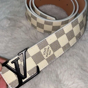 Rutig Louis Vuitton skärp med LV-spänne - Snyggt skärp från Louis Vuitton med klassiskt rutigt mönster i beige och grått. Skärpet har en stor svart LV-logga som spänne och är gjort i skinn med vita kanter. Perfekt för att lyfta vilken outfit som helst. 