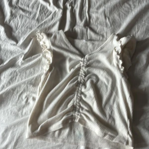 Vit linnetopp med volangärmar - Supersöt vit linnetopp med volangdetaljer på axlarna och snygg rynkning framtill. Toppen har v-ringning och är ärmlös, perfekt för varma dagar. Materialet är lätt och har en diskret struktur som ger en extra touch.