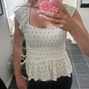 Vit blus med volang och knyt - Supersöt vit blus med små mörkblå prickar, volangärmar och smockad överdel. Toppen har en söt knytning i nacken och lätt utställd nederdel som ger en flowy känsla. Perfekt för sommarens alla tillfällen!