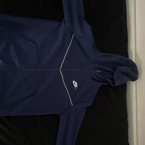 Blå Nike tech med huva - Säljer en marinblå Nike tech i sportig stil med huva och hel dragkedja. Jackan har vita detaljer och logga på bröstet, samt två stora fickor framtill. 