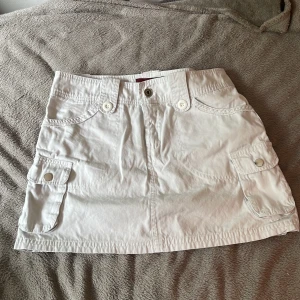 Vit kortkjol från Tiger King - KOLLA MIN ANDRA ANNONS FÖR EN BRA DEAL SOM INKLUDERAR DENNA KJOL!! Vit kortkjol från Tiger King med stora fickor på sidorna och snygga knappdetaljer både fram och bak. Kjolen har bälteshällor och är gjord i bomull, perfekt för en somrig y2k look.