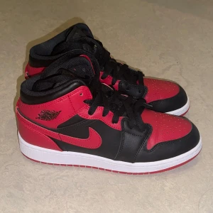 Nike Air Jordan 1 Mid Svart/Röd - Finns ingen box. Har knappt använt dom. Sist jag använde var några år sen och har inte haft på mig sen dess.                     Snygga Nike Air Jordan 1 Mid sneakers i klassisk svart och röd färg med vita sulor. Skorna har svart snörning, röd swoosh och ikonisk Air Jordan-logga på sidan. Tillverkade i skinn och syntet, med rund tå och platt sula. Perfekta för dig som vill sticka ut med streetstyle.  