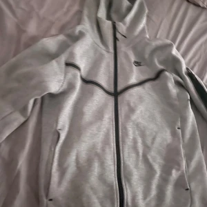 Nike tech grå - Snygg grå hoodie från Nike med hel dragkedja och svarta detaljer. Tröjan har huva och långa ärmar, perfekt för en sportig look eller chill dagar. Klassisk Nike-logga på bröstet och bekväm passform.