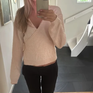 Beige omlott kofta med knyt - Mysig rosa omlott kofta med knyt i sidan och v-ringad hals. Tröjan är stickad och har långa ärmar samt ribbade muddar. Perfekt för lager-på-lager och en chill look.