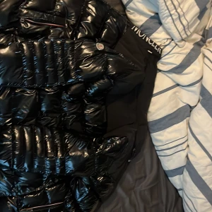 Svart pufferjacka från Moncler - Säljer en svart, glansig pufferjacka från Moncler med klassisk logga på ärmen. Jackan har quiltad design, dragkedja och hög krage. Perfekt för kalla dagar och riktigt snygg streetstil. Fodrad och med flera fickor för extra funktionalitet. Jackan är storlek M