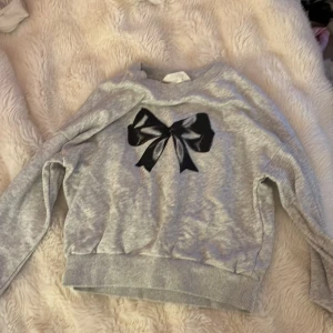 Grå sweatshirt med svart rosett H&M - Grå sweatshirt från H&M med en stor svart rosett framtill. Tröjan har rund hals, långa ärmar och ribbade muddar vid ärmslut och nederkant. Perfekt för dig som gillar en söt och stilren look.