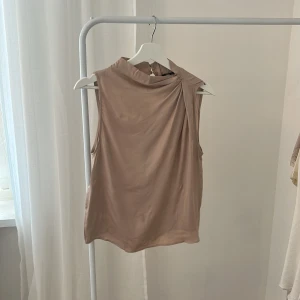 Beige satin topp från Zara - Säljer en stilren beige blus i mjukt, glansigt material. Toppen är ärmlös med hög krage och har en snygg drapering vid axeln. Stängs med knapp i nacken och har en lätt flowy passform. Perfekt för dig som gillar minimalistisk och chic stil.