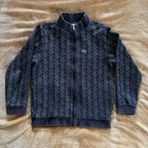 Svart mönstrad zipjacka från Lacoste stl.140 - Svart zipjacka från Lacoste med grått geometriskt mönster över hela jackan. Klassisk ståkrage, hel dragkedja framtill och broderad Lacoste-logga på bröstet. Ribbstickade muddar vid ärmslut och nederkant. Perfekt för en chill och sportig look. Stl.140 nypris ca 1400:- köpt i våras på Kids brandstore.