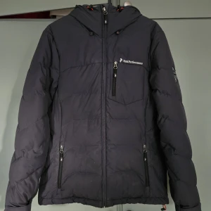 Svart Peak Performance dunjacka - Säljer en svart dunjacka från Peak Performance med huva och flera dragkedjeförsedda fickor. Jackan har logga på bröstet och ett märke på ärmen. Perfekt för kalla dagar och har en stilren, sportig look. Färg: mörkgrå