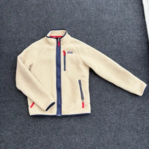 Beige fleecejacka från Patagonia - Säljer en beige fleecejacka från Patagonia med marinblå detaljer och röda dragkedjor. Jackan har hög krage, hel dragkedja framtill och tre fickor med dragkedja. Perfekt för hösten/vinter och riktigt skön att ha på sig. Storlek 164/XL i barn storlek.