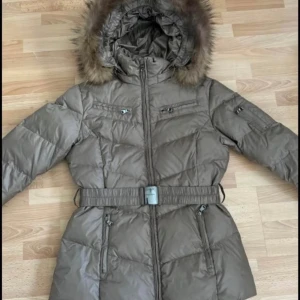 Jacka med päls och bälte - Snygg jacka med avtagbar huva och päls. Jackan har bälte i midjan, flera dragkedjefickor och quiltad design. Perfekt för kalla dagar med sin långa modell och varma material. Stängs med dragkedja och har långa ärmar. Storlek 38, bröst 100cm, längd 70cm, färg khaki/brun.