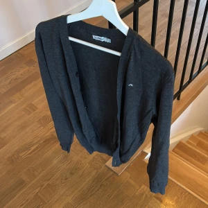 Cardigan  J.Lindeberg - Snygg mörkgrå kofta från J.Lindeberg med diskret logga på bröstet. Koftan har v-ringning, knappar framtill och långa ärmar med ribbade muddar. Tillverkad i mjuk bomull, perfekt för lager-på-lager och chill stil.