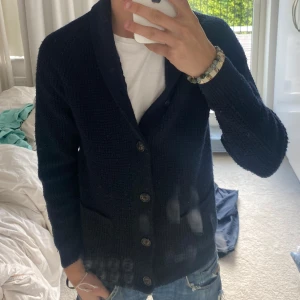 Mörkblå stickad kofta - Säljer en mörkblå stickad kofta med grov struktur och snygg krage. Koftan har långa ärmar, knappar framtill och två praktiska fickor. Perfekt för dig som gillar klassisk och chill stil.