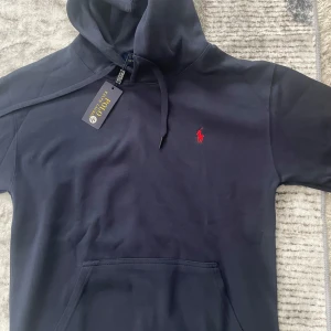 Mörkblå hoodie från Polo Ralph Lauren - Snygg mörkblå hoodie från Polo Ralph Lauren med klassisk röd logga broderad på bröstet. Tröjan har huva med snörning, känguruficka och långa ärmar. Tillverkad i mjuk bomull som är perfekt för chill dagar.