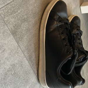 Svarta sneakers med vit sula - Säljer ett par svarta sneakers med vit platt sula och rund tå. Skorna har snörning och är tillverkade i skinn med textilfoder. Stilrena och passar perfekt till jeans eller shorts. Klassisk design med diskreta sömmar och svart ovandel. 