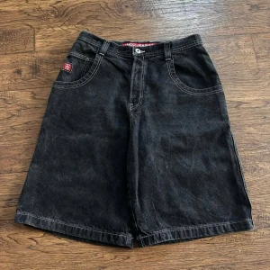 Svarta JNCO Jeans shorts med brodyr - Riktigt feta svarta jeansshorts från JNCO Jeans med bred passform och kontrastsömmar. På baksidan finns en stor broderad tiger på fickan och röda JNCO-loggor. Shortsen har klassisk femficksdesign och är tillverkade i robust denim. Perfekt för dig som gillar streetwear och vill sticka ut.