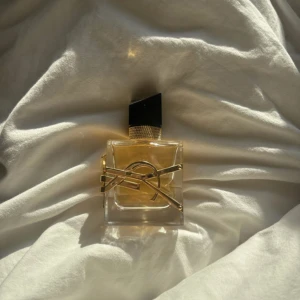 Yves Saint Laurent Libre Eau de Parfum - Säljer en ikonisk YSL Libre parfym i snygg glasflaska med guldiga detaljer och svart asymmetrisk kork. Flaskan har en lyxig känsla med YSL-loggan i metall runt flaskan och gyllene vätska inuti. Perfekt för dig som gillar eleganta och trendiga dofter.75% kvar 