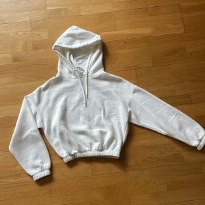 Vit croppad hoodie från Bershka - Säljer en vit croppad hoodie från Bershka med huva och snörning. Tröjan har långa ärmar och resår i midjan och vid ärmsluten för en snygg passform. Perfekt för en chill och stilren look.