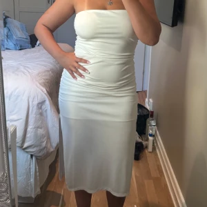 Vit axelbandslös långklänning - Säljer en vit axelbandslös långklänning med snygg rynkning längs sidorna och en slits bak. Klänningen har en tight passform och är gjord i ett mjukt, stretchigt material som sitter skönt mot kroppen. Perfekt för dig som gillar minimalistisk och stilren look. Den är använd så lite nopprig och någon fläck men inget som man tänker på