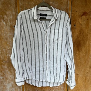 Randig linneskjorta från Nano Universe - Snygg vit skjorta i linne från Nano Universe med svarta och gråa vertikala ränder. Skjortan har klassisk krage, bröstficka och knäppning framtill. Perfekt för en chill och stilren look under varmare dagar.