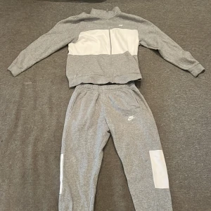 Grå träningsväst från Nike M - Nike väst i grått med vit panel framtill, dragkedja hela vägen och ståkrage. Västen har en sportig look och är gjord i mjuk bomullsmix som känns skön mot kroppen. Perfekt för gymmet eller chill dagar. Snyggt broderad Nike-logga på bröstet. Hela setet för 399kr