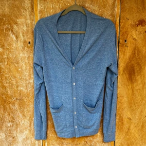 Blå kofta från American Apparel M - Snygg blå kofta från American Apparel i storlek M. Koftan har v-ringning, knäppning framtill och två praktiska fickor. Tillverkad i en mjuk mix av polyester, bomull och viskos. Perfekt för lager-på-lager och chill vardagsstil.