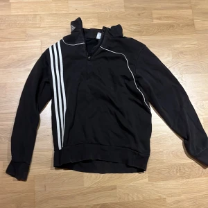 Svart Adidas half zip tröja - Svart långärmad Adidas-tröja med klassiska vita ränder på ena sidan och vit piping. Tröjan har en hög krage med halv dragkedja och Adidas-logga broderad på kragen. Perfekt för sportiga vibes eller chill dagar.