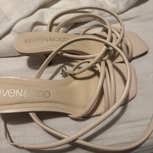 Beige sandaler med smala remmar Even&Odd - Snygga beige sandaler från Even&Odd med smala remmar som slingrar sig runt foten och ankeln. Skorna har fyrkantig tå, låg klack och spänne vid ankeln. Tillverkade i syntetmaterial och perfekta för en trendig look.
