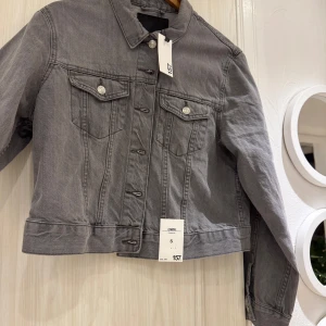 Grå jeansjacka, storlek S - Snygg grå jeansjacka från lager 157i klassisk modell med knappar framtill och två bröstfickor med lock. Jackan har lång ärm och raka linjer, perfekt att slänga på sig över en hoodie eller t-shirt. Enkel och stilren design som funkar till det mesta.