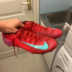 Röda Nike Mercurial fotbollsskor - Snygga röda Nike Mercurial fotbollsskor med turkosa detaljer och logga. Skorna har snörning, platt sula med dobbar och är tillverkade i syntetmaterial. Perfekta för dig som vill sticka ut på planen med en sportig och modern look.