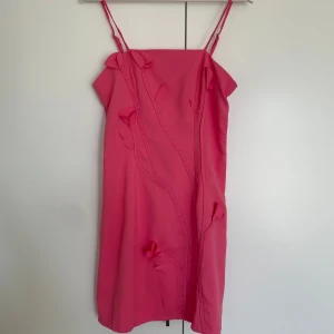 Rosa kortklänning med blommiga detaljer - Säljer en superfin rosa kortklänning med smala axelband och 3D-blommor som dekoration framtill. Klänningen har en rak passform och är gjord i ett mjukt syntetmaterial. Perfekt för dig som vill sticka ut med coola detaljer och en härlig färg.