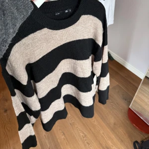 Randig stickad tröja  - Säljer en stickad tröja med vida armar. Tröjan har breda svarta och beige ränder, rund halsringning och långa ärmar. 