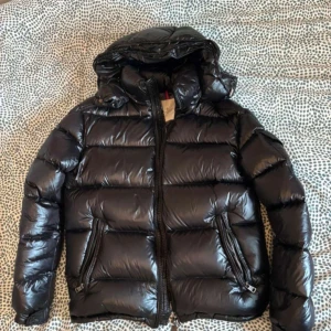 Moncler jacka - Säljer en svart, glansig dunjacka från Moncler med huva och dragkedja framtill. Jackan har två snedställda fickor med dragkedja och klassisk Moncler-logga på ärmen. Perfekt för kalla dagar och riktigt snygg streetstil.