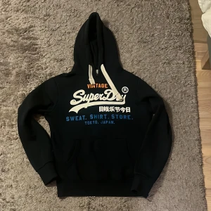 Mörklblå Superdry hoodie med tryck - Mörkblå hoodie från Superdry med tryck på bröstet. Tröjan har huva med vita snören, känguruficka och mjukt foder. Perfekt för dig som gillar streetwear och vill ha en snygg och bekväm look.