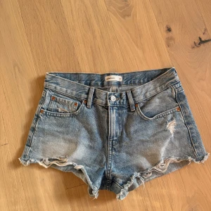 Blå jeansshorts från Gina Tricot - Snygga blå jeansshorts från Gina Tricot i klassisk femficksmodell. De har råa och fransiga kanter samt slitningar framtill för en cool vibe. Materialet är robust denim och passformen är normal. Perfekta för varma sommardagar!