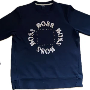 Svart sweatshirt från Hugo Boss - Riktigt fet Svart sweatshirt från Hugo Boss med stort BOSS-tryck i vitt och beige på bröstet. Tröjan har rund halsringning, långa ärmar och är tillverkad i mjuk bomull. Perfekt för en stilren och avslappnad look.