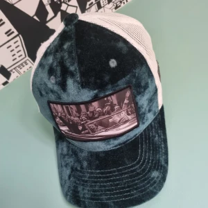 Mörkgrön sammetstruckerkeps med tryck - Säljer en unik truckerkeps i mörkgrön sammet med vit mesh på sidorna och bak. Framtill finns ett stort svartvitt tryck med motiv av människor i en bil. Kepsen har böjd skärm och cool vintagekänsla, perfekt för dig som vill sticka ut.