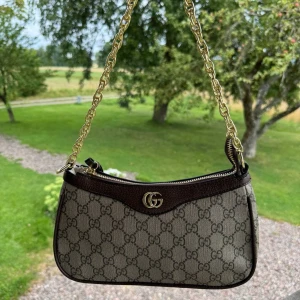 Gucci axelväska med GG-mönster - Snygg axelväska från Gucci med klassiskt GG-mönster i beige och brunt. Väskan har guldfärgad kedja som axelrem, bruna skinn-detaljer och dragkedja upptill. Insidan är fodrad med ljus textil och har präglad Gucci-logga på skinnet. Perfekt storlek för det viktigaste.