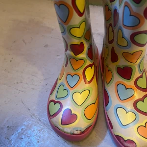 Färgglada hjärt-mönstrade stövlar - Snygga gummistövlar med färgglatt hjärtmönster i rött, gult, grönt och blått. Stövlarna har rosa detaljer och sula, samt mjukt foder på insidan. Perfekta för regniga dagar när du vill sticka ut med din stil.