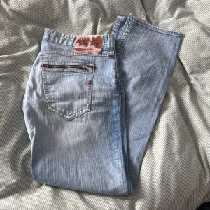 Ljusblå Replay jeans, w33 l32 - Säljer ett par ljusblå jeans från Replay med klassisk femficksdesign och snygga detaljer som dragkedja på bakfickan och gul Replay-brodyr. Jeansen är raka i modellen och tillverkade i bomull. Perfekta för en avslappnad och trendig look.