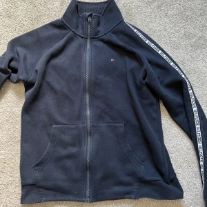Svart ziptröja Tommy Hilfiger - Svart ziptröja från Tommy Hilfiger med logoband längs ärmarna och liten broderad flagga på bröstet. Hög krage, två fickor fram och hel dragkedja. Skön passform och klassisk streetstil, perfekt till jeans eller joggers. Det är lite småhål på ryggen men har man en svart t-shirt under så syns det inte lina mycket. Nypris- 999