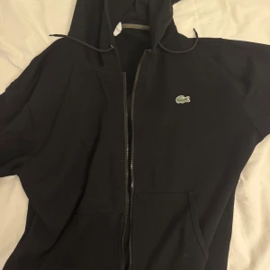 Svart hoodie från Lacoste med dragkedja - Svart hoodie från Lacoste i klassisk passform med dragkedja framtill och huva med snörning. Tröjan har fickor på magen och den ikoniska krokodilloggan på bröstet. Perfekt för en chill och stilren look.