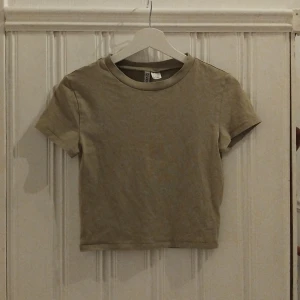 Beige croppad topp från Divided - En enkel och stilren croppad topp från Divided i beige färg. T-shirten har rund halsringning och korta ärmar, perfekt att matcha med jeans eller kjol. Tillverkad i mjuk bomull för en skön känsla hela dagen.