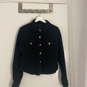 Svart manchester overshirt med knappar - Snygg svart overshirt i manchester från SheIn med silvriga knappar framtill och två stora bröstfickor. Jackan har en boxig, croppad passform och klassisk krage. Perfekt att slänga över en hoodie eller t-shirt för en chill look. (Aldrig använd)