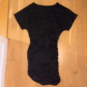 Svart kort klänning med knyt - Säljer en svart kort klänning med snygg knytning i midjan och rynkade detaljer längs sidorna. Klänningen har korta ärmar och en mjuk, följsam passform som framhäver siluetten. Perfekt för dig som gillar stilrena och enkla plagg med en twist.