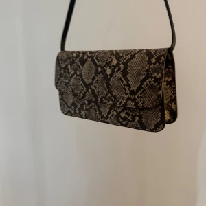 Axelväska med ormmönster i skinnimitation - Cool axelväska i svart och beige med trendigt ormmönster. Väskan har en stilren, rektangulär form och smal axelrem. Köpt från ASOS🤎