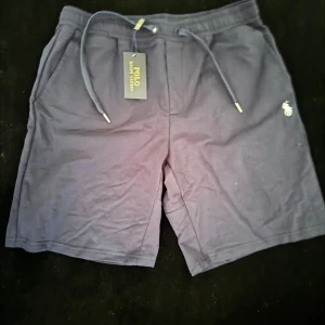 Mörkblå shorts från Polo Ralph Lauren - Snygga mörkblå shorts från Polo Ralph Lauren med klassisk logga broderad i vitt på benet. Modellen har snörning i midjan och är tillverkad i mjuk bomull. Perfekta för chill dagar och har en normal passform med praktiska fickor. Skriv om du behlver mer bilder/ detaljer. ALDRIG ANVÄNT INNAN
